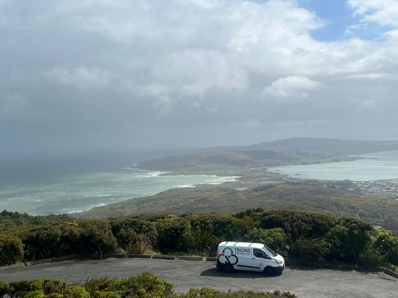 Trions Electrical van parked on a ridge overlooking a wide coastal landscape under a cloudy sky.
